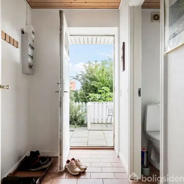 Dør står åben i en flisebelagt entré med sko på gulvet. Udenfor ses en terrasse og grøn beplantning. Til højre er en dør til et badeværelse med toilet.