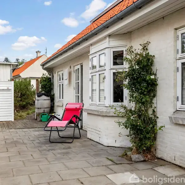 To pink liggestole står på en flisebelagt terrasse foran et hvidt murstenshus med rødt tegltag. Ved siden af gror en grøn plante langs væggen.