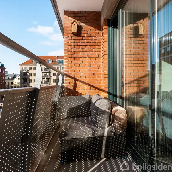 En altan med en grå stol står op ad en murstensvæg. Der er glasdøre til højre og et udsyn til bybygninger i baggrunden under en blå himmel.