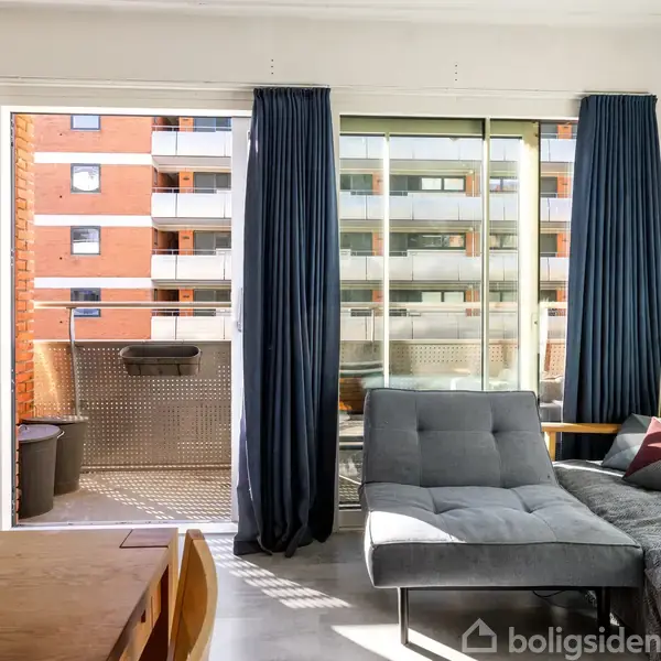 Sofa med puder står i en lys stue tæt på en glasdør, der fører ud til en altan med udsigt over en rød murstensbygning. Mørke gardiner hænger ved døren.