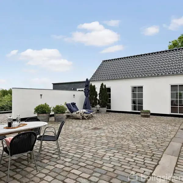 Terrasse med flisebelægning og møbler, herunder et spisebord og stole. Solsenge og parasol står i nærheden. Hvide bygninger med sorte tegltage omgiver området. Blomsterkrukker dekorerer omgivelserne.