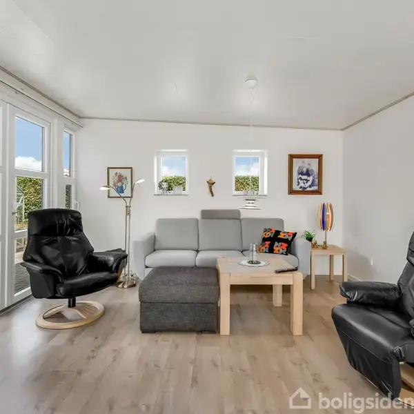 Sofa og lænestole står afslappet i en lys stue med trægulv. Store vinduer åbner mod en terrasse og have. Kunstneriske værker pryder de hvide vægge, og et lille sofabord er i centrum.