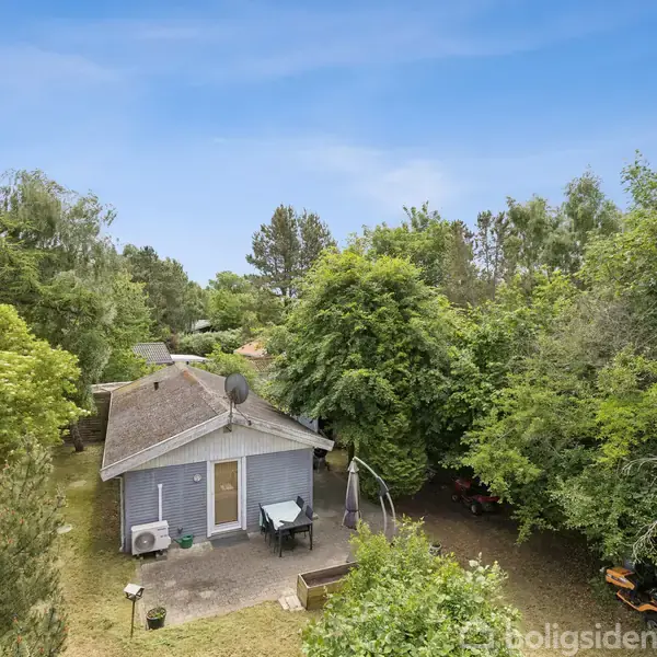 Et lille hus står omgivet af tæt skov og grønne træer, med en terrasse med borde og stole foran huset under en klar blå himmel.