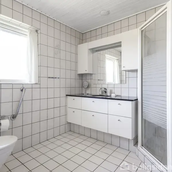 Et rummeligt badeværelse har hvide fliser og lyst indretning. En håndvask med spejlskab og skuffer er placeret ved væggen. Der er også en brusekabine og toilet i rummet.