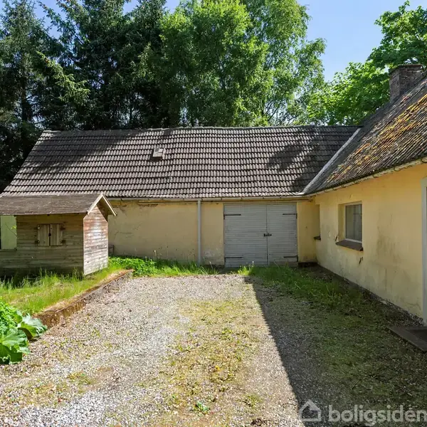 Et gammelt bondehus med en gul facade står tomt. En grusvej fører til en garage med dobbelt dør. Tætte træer omgiver området, der er præget af natur og ro.