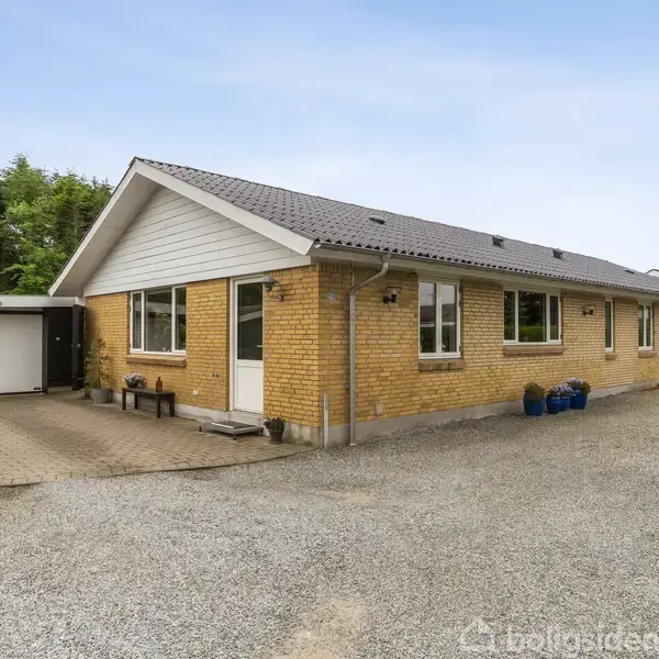 En gul murstensbygning med gråt tag står stille. En grusvej fører op til en hvid garage. Omgivet af grønne buske og træer.