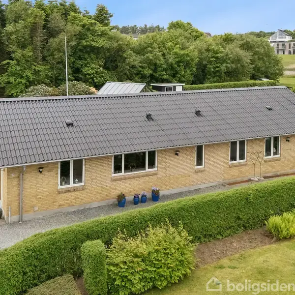En gul murstensbygning står stille i en frodig have med klippede buske. Den har et mørkt tag og flere vinduer. Bagved ses et grønt område med træer og moderne bygninger.