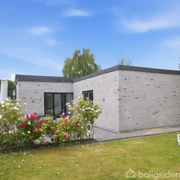 En moderne, enkeltrums bolig med grå murstensfacade står i en velplejet have. Omringet af blomstrende buske, har det flade tag store vinduer. Et hvidt havebord med stole står i græsset.