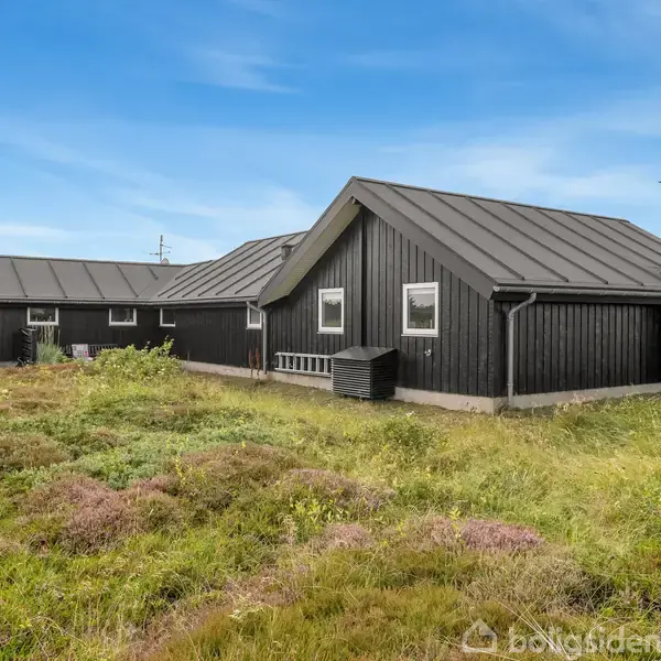 Sort træterrasse med bord og stole står tom ved siden af en sortmalet bygning med store vinduer. Omgivet af grønne buske og græs i et sommerhusområde.