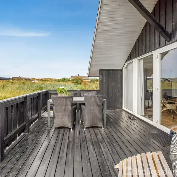 Sort træhus med gråt tag står stille omgivet af lav vegetation og græs. Husnummer 46 ses tydeligt. Himlen er klar og blå i baggrunden.