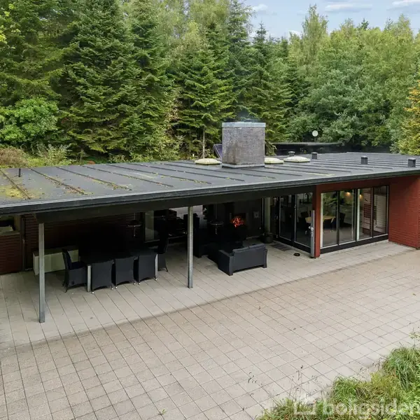 En moderne, flad taghustype omgivet af skov. Foran huset er en stor terrasse med havemøbler. Bagsiden af huset har store glaspartier, der åbner mod skovens tætte grønne bevoksning.