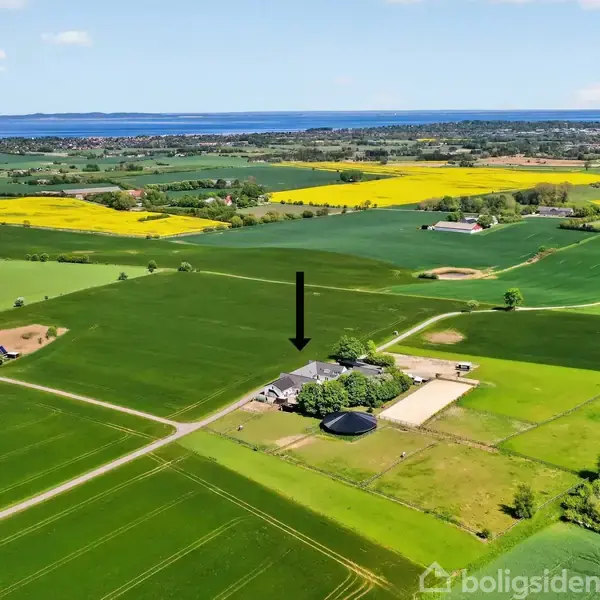 Landejendom markeret med en pil, omgivet af store grønne marker. Fjern udsigt viser havet og spredt bebyggelse i det bakkede landskab. Skyfri himmel i baggrunden.