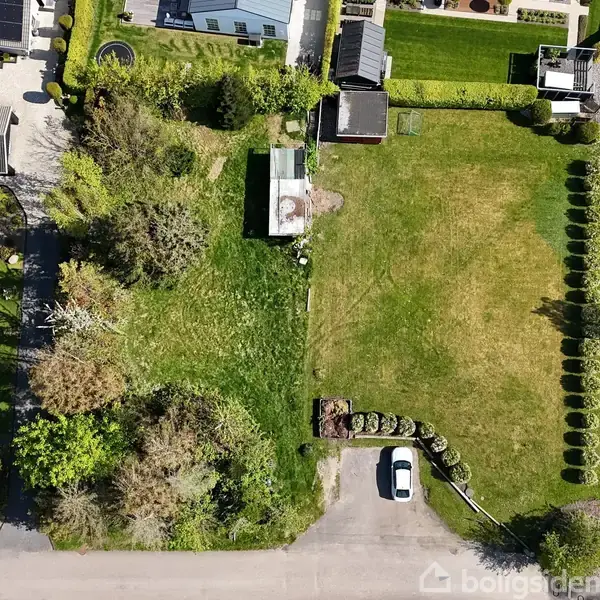 Græsplæne med buske og træer, omgivet af huse og haver. En bil er parkeret ved vejen i forgrunden. Beplantning langs kanterne skaber symmetri.