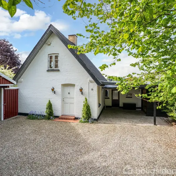 Hvidt hus med stejlt tag og skorsten står på grusbelagt gårdsplads. Carport til venstre, omringet af grønne træer under en blå himmel med skyer.