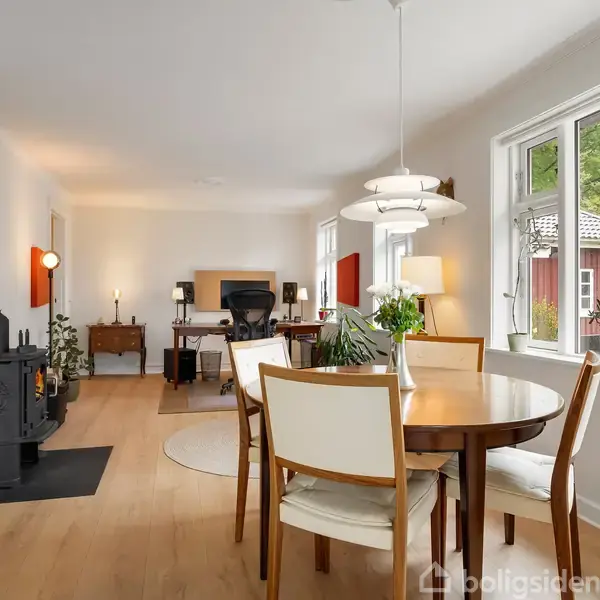 Spisebord med stole står i en lys stue med trægulv. En brændeovn er placeret ved væggen, mens et vindue viser en udsigt til en rød bygning og grønne træer udenfor.