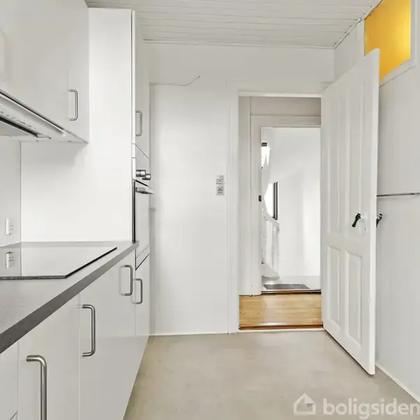 Køkken med hvide skabe på begge sider, sort bordplade, åben dør i enden fører til en lys gang. Indbygget ovn og et køleskab fra Whirlpool ses.