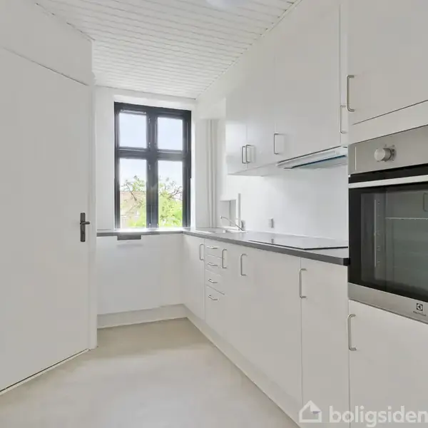 Et hvidt køkken har moderne skabe og en indbygget ovn. Rummet er lyst op af et stort vindue, der giver udsigt til grønne områder udenfor.