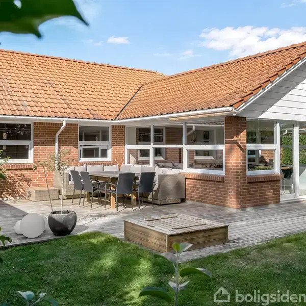 Et rødt murstenshus med rødt tegltag har en terrasse med bord og stole. Det ligger i en grøn have omkranset af vegetation under en blå himmel.