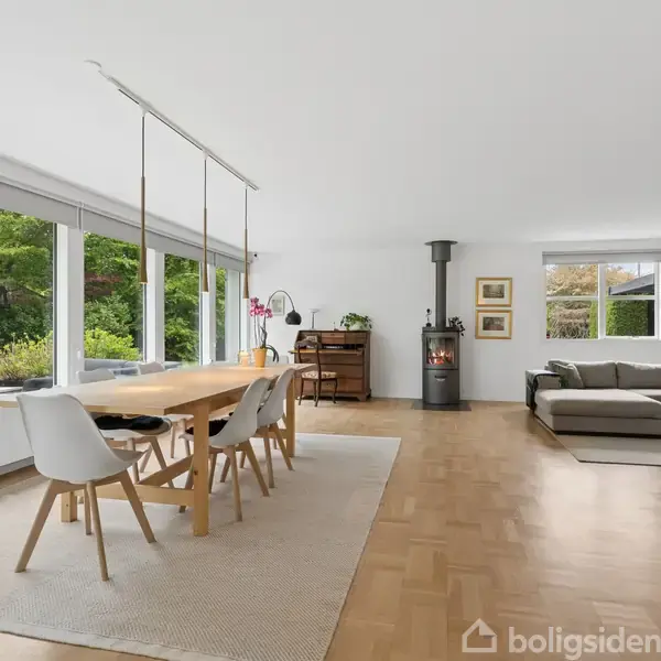 Et moderne spisestueområde med et stort træbord og stole, der står på et lyst gulvtæppe. Store vinduer slipper naturligt lys ind. I baggrunden findes en brændeovn og sofa.