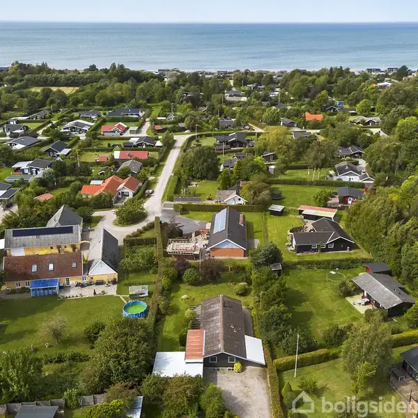 Luftfoto af en villaby med mange huse, hvor nogle har røde tage, omgivet af grønne haver. I baggrunden ses skov og havet, der strækker sig mod horisonten.