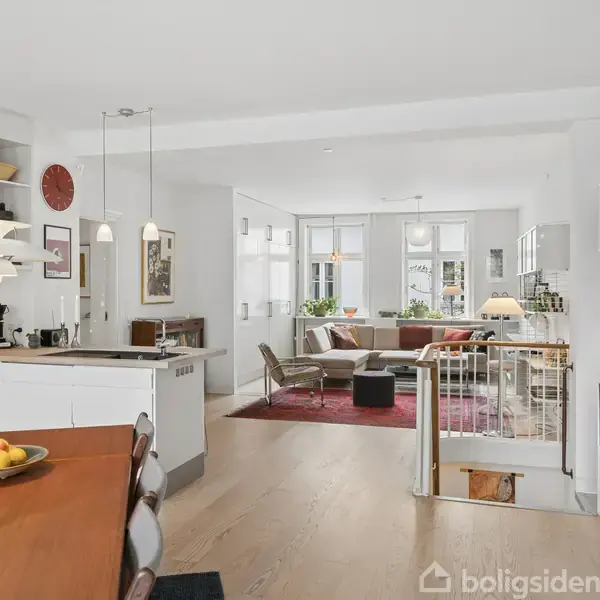 Lyst køkken med spisebord, moderne hvide skabe og apparater. Soveværelse med dobbeltseng mod en accentvæg. Stue med stor hjørnesofa, sofabord og TV, lyse vægge og gulv.