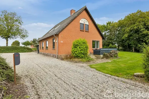 En rød murstensbygning med tagvinduer står ved en grusbelagt indkørsel. Nummer 44 er synligt på muren. En velholdt græsplæne og træer omkranser huset. Skiltet viser "Elmely Ø. Knudstrup".