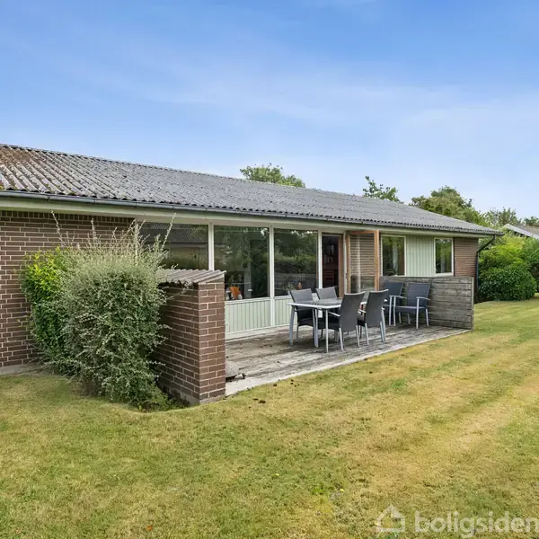 Et enkelt etplanshus i mursten med store vinduer står på en velplejet græsplæne. Foran huset er der en terrasse med havemøbler, omgivet af træer og buske.