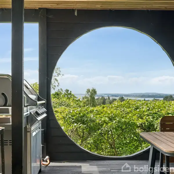 En træterrasse med en grill og et bord med vinglas. En stor cirkulær åbning giver udsigt til grønt landskab og himmel. Vinglas og natur skaber en fredelig atmosfære.