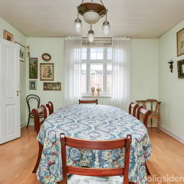 En ovalt spisebord med blåmønstret dug omgivet af seks stole. Rummet har lyse vægge, trægulv, en åben dør til køkkenet, vægdekorationer og en hængende loftslampe.