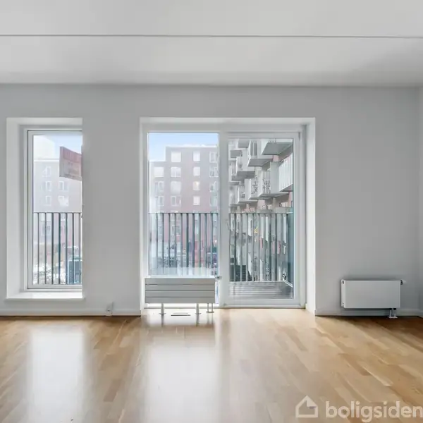 Lyse trægulve pryder en rummelig stue med to høje vinduer og en glasdør. Udsigt til en moderne bygning gennem vinduerne med en radiator på højre side.