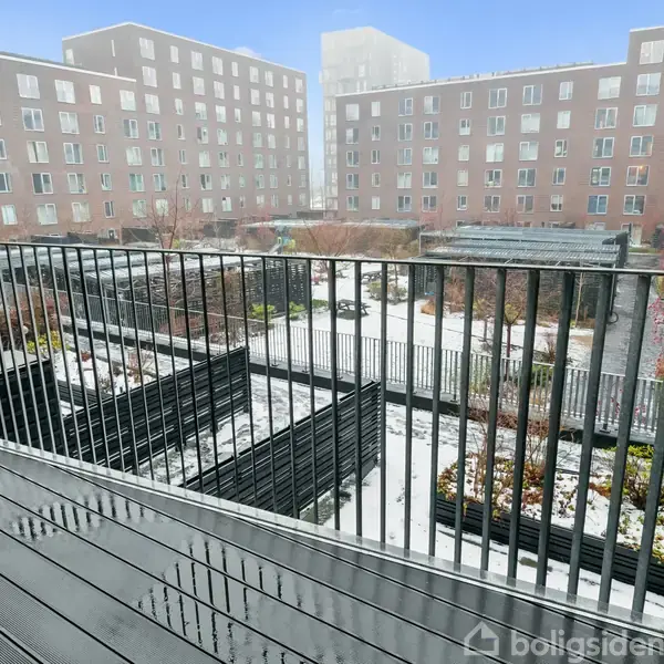 Balkon med metalgelænder, udsigt til sneklædt gårdområde med plantekasser og moderne lejlighedsbygninger i baggrunden. Klare vinterlige omgivelser under en blå himmel.