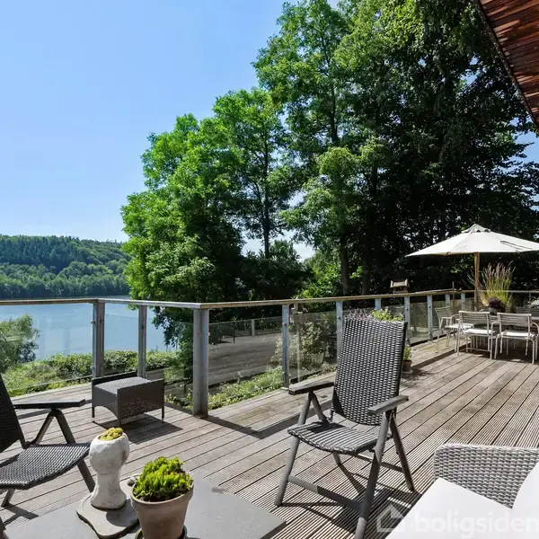 Terrasse med sorte stole og et bord, der kigger ud over en sø omgivet af grønne træer. Sollys skinner igennem på området med parasol og udendørs spisebord.