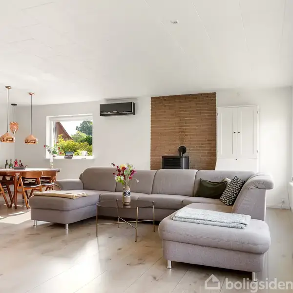 Gråt hjørnesofa står i en lys stue med træplanker, placeret ved vinduer. I baggrunden ses spisebord med stole og kobberlamper. Væggen har en brændeovn og skab.