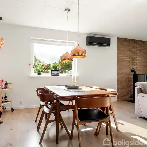 Spisebord med træstole står i en lys stue. Kobberlamper hænger over bordet, blomster dekorerer et skab, og der er et klimaanlæg og en sofa ved murstensvæggen.