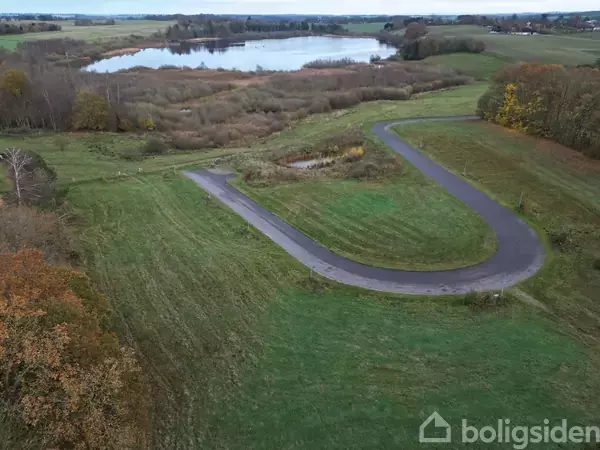 Asfalteret vej svinger gennem grønt landskab, omgivet af træer og græsarealer, med en sø i baggrunden under en skyfri himmel.