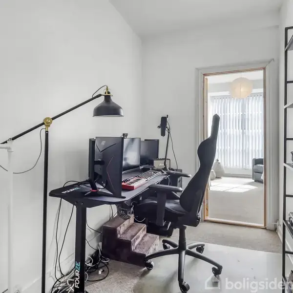 En kontorstol står foran et skrivebord med to computerskærme. Rummet har en minimalistisk indretning, og en åben dør fører til et lyst værelse med en sofa i baggrunden.