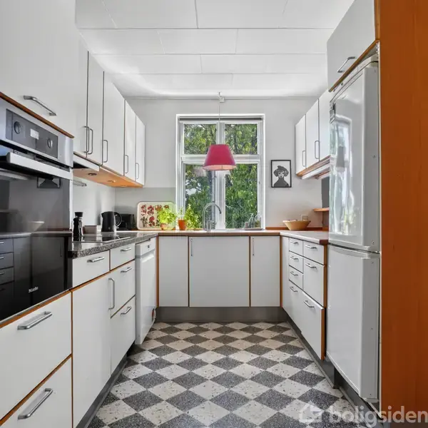 Et lyst køkken med hvidt interiør og trækanter. En rød lampe hænger over vasken, som står ved et vindue med udsigt til grønne træer. En vægdekoration ses til højre.