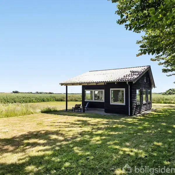 Et sort træhus med store vinduer står roligt på en åben græsplæne omgivet af frodig vegetation og åbne marker under en klar blå himmel.