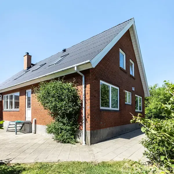 Rødt murstenshus med skråt tag står på et flisebelagt terrasseområde omgivet af grønne buske under en klar blå himmel. Bord og stole står ved husets gavl.