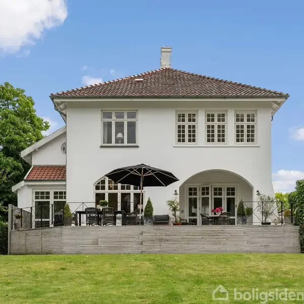 Hvid villa med rødt tegltag står på en grøn græsplæne. Terrassen har havemøbler og en stor parasol. Skyfri blå himmel og træer omkranser huset.
