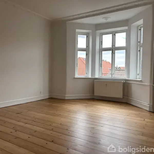 Et tomt værelse med trægulv og hvide vægge. Vinduet i karnappen giver udsigt til hustage og himmel. Radiator placeret under vinduet.
