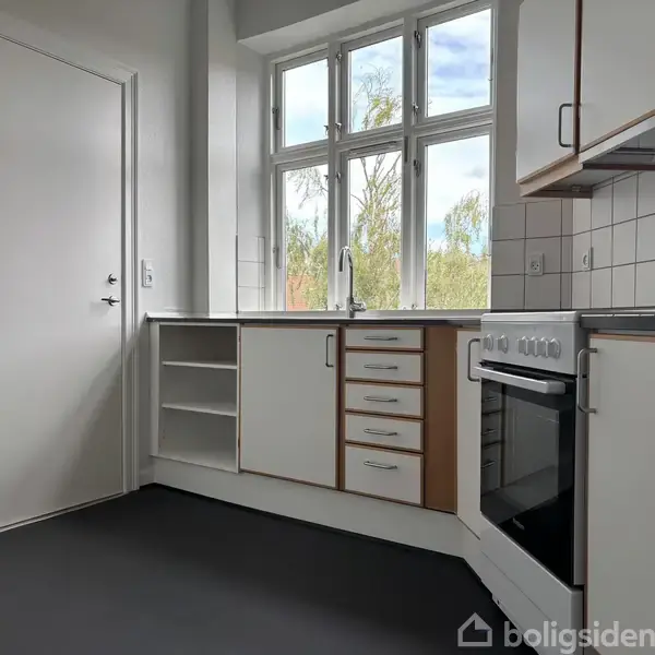 Et funktionelt køkken med hvide skabe og skuffer. Der står en ovn med kogeplade ved siden af en vask. Et stort vindue giver udsigt til træer udenfor.
