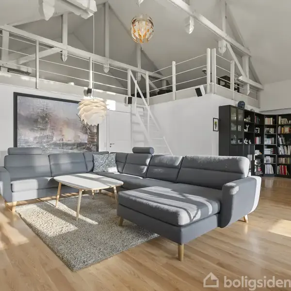 Gråt hjørnesofa-arrangement står på et lyst trægulv. En lille trappe fører til en højtliggende hems. Store reoler fyldt med bøger udgør noget af væggen i baggrunden.