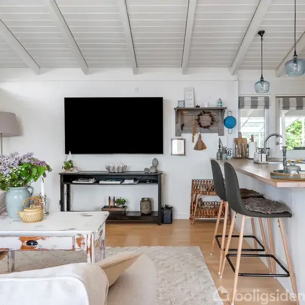 En hyggelig stue med sofa og sofabord med blomster på. Et stort TV hænger på væggen. En åben køkkenbar med høje stole og dekorativ belysning ved siden af.