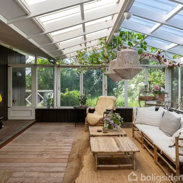 Loungesæt bestående af sofa, bord og stol står i en lys udestue med glasvægge og -tag. Grønne planter hænger fra loftet, og udsigten viser frodig have udenfor.