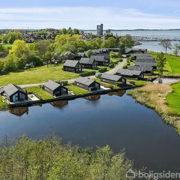 Flere moderne træhuse står langs en lille sø; omgivet af grønne marker og træer, med en fjord og en by set i baggrunden.