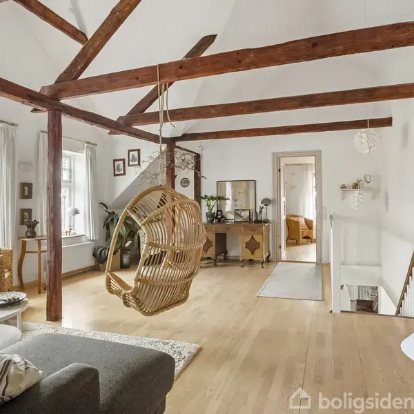 En hængestol af flet svæver i et rummeligt, lyst stueområde med synlige træbjælker. Rummet har trægulv, sofa, og store vinduer, der skaber en indbydende atmosfære.