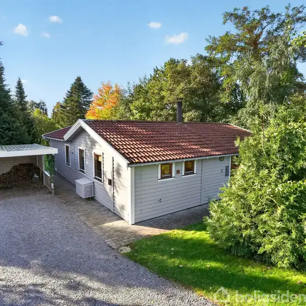 Et enfamiliehus med rødt tegltag står i en grøn have. En grusbelagt indkørsel fører til en carport til venstre. Store træer omgiver ejendommen.