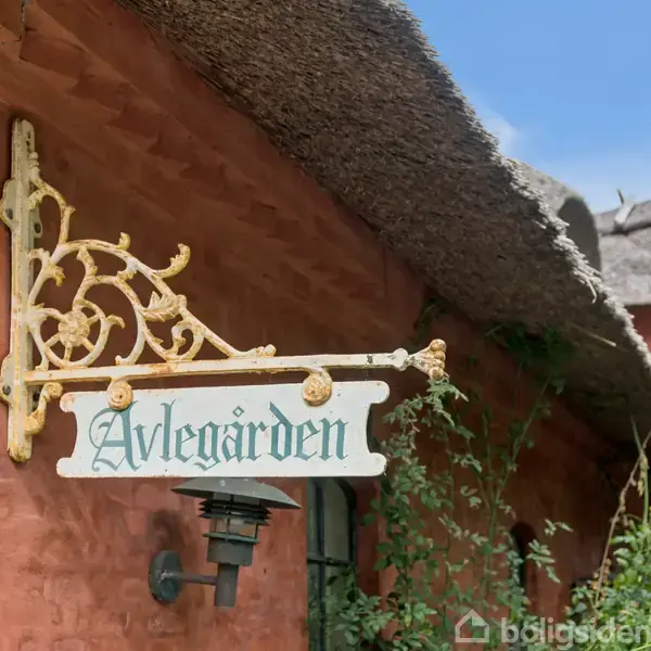 Et skilt med teksten "Avlegården" hænger på en detaljeret metalholder. Skiltet er monteret på en rød bygning med stråtag, omgivet af grøn vegetation og blå himmel.