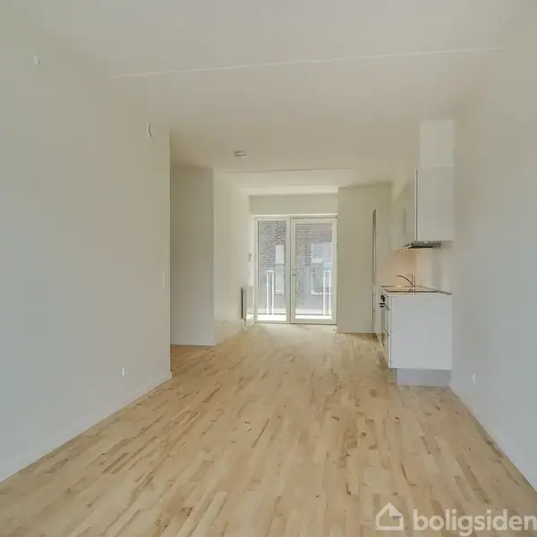 Rummeligt, lyst værelse med trægulv og en lille integreret køkkenniche. I baggrunden er der store glasdøre, der fører ud til en altan. Hvide vægge og loft.